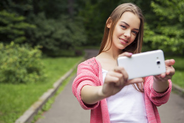 Beautiful brunette woman in neutral casual outfit walking in park, make selfie using her phone, smiling. Lifestyle portrait. Copy space