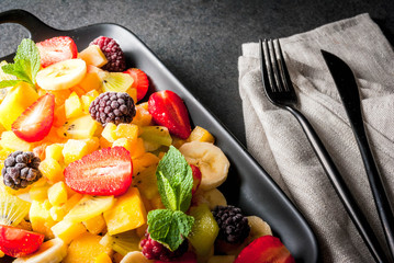 Vegan dietary foods. Vitamins. Dessert. Summer. Salad of fresh organic fruits mango, peach, apple, banana, kiwi, strawberry, blackberries. On black ceramic plate, black stone table copy space