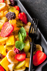 Vegan dietary foods. Vitamins. Dessert. Summer. Salad of fresh organic fruits mango, peach, apple, banana, kiwi, strawberry, blackberries. On black ceramic plate, black stone table copy space