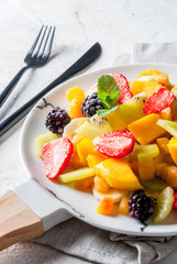 Vegan dietary food. Vitamins Summer dessert. Salad of fresh organic fruits mango, peach, apple, banana, kiwi, strawberry, blackberries. On white marble plate, white concrete table copy space