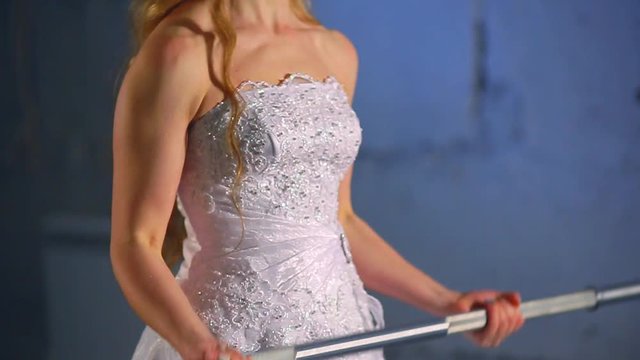 Beautiful and muscular bride blonde in a wedding dress doing an exercise with a barbell in a gym, crossfit