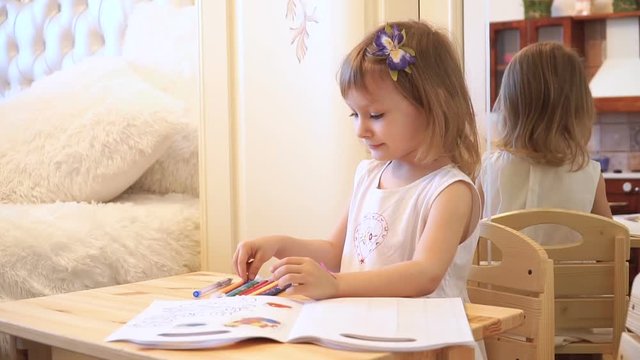Active little preschool age child, cute toddler girl with blonde curly hair, drawing picture on paper using colorful pencils and felt-tip pens, sitting at wooden table indoors at home or kindergarten