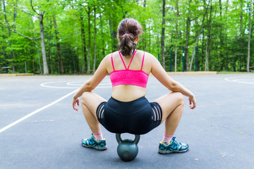 Back of young fit woman sitting on kettlebell in outdoor park in workout clothes during summer