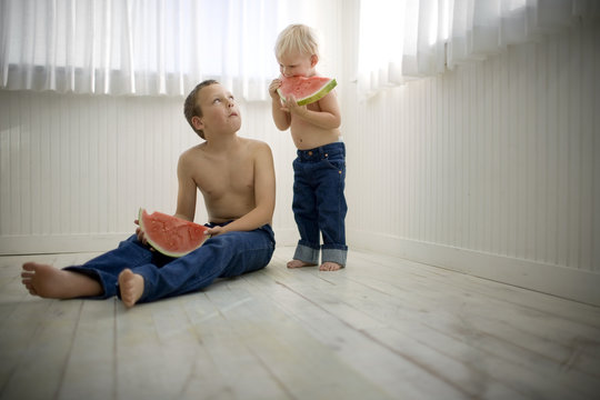 Two shirtless brothers eating slice of watermelon inside a bare room.