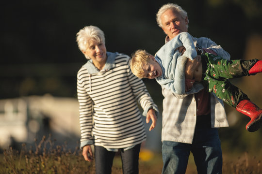 Small Boy Laughs As His Grandfather Playfully Hefts Him Up Horizontally And His Grandmother Smiles As She Watches While They Walk Together Outside.