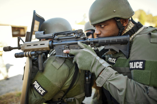 Male police officer aims a gun toward a target as another police officer holds up shield.