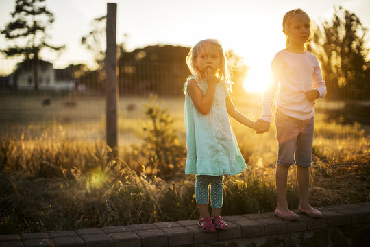 Two young blond sisters hold hands at the side of a country road at sunset.