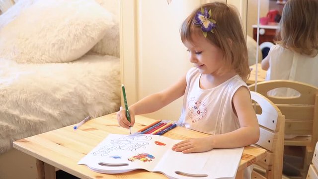 Active little preschool age child, cute toddler girl with blonde curly hair, drawing picture on paper using colorful pencils and felt-tip pens, sitting at wooden table indoors at home or kindergarten