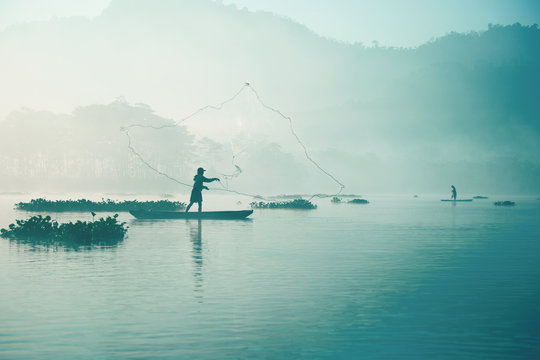 Fisherman casting out his fishing net in the river by throwing it high up into the air early in the blue colored morning to catch fish with his little fishing boat.