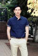 Молодой красивый брюнет мужчина позирует в синей рубашке, светлых летних штанах и часах
