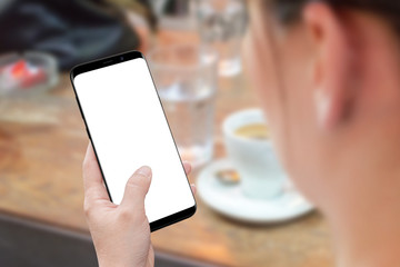 Woman holding modern smart phone with isolated screen for mockup. View behind shoulder. Desk with coffee in background.