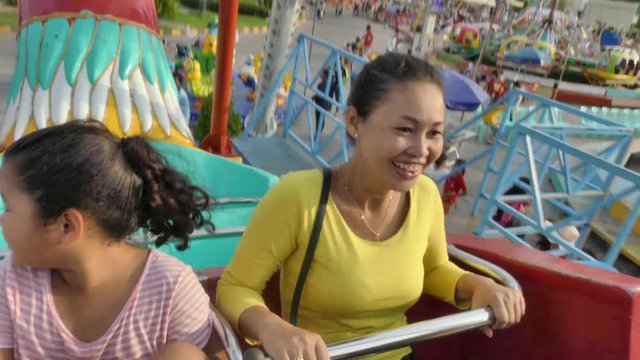 A multicultural Asian family rides on an amusement park swing ride together; shot moves from mother and daughter to settle on son, while other rides can be seen below 