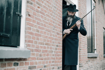 Man with long beard dark dressed standing with rifle ready to shoot.