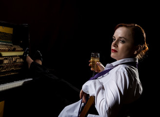 Elegant woman in a white shirt and tie sitting next to the piano and drinks champagne
