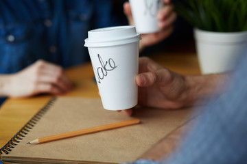 Closeup shot of two paper coffee cups with names on them in hands of young people working in cafe