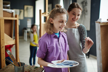Portrait of happy boy enjoying painting class in art studio, with young teacher helping him at easel