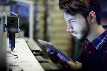 Side view portrait of modern factory worker using digital tablet to operate laser cutting machine...
