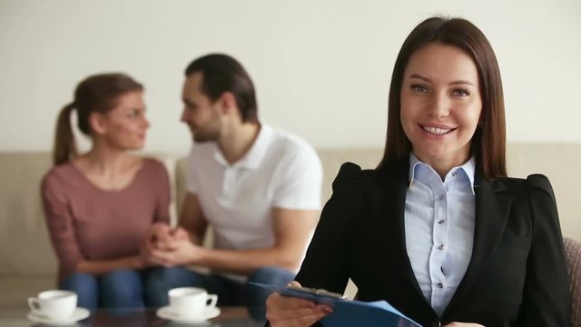 Portrait of professional female relationship therapist sitting in her office after therapy session looking at camera with confident expression, young happy reconciled couple on the background