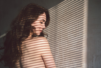 Obraz premium Side view of a beautiful young woman standing near the wall at home a with bare shoulders and have shadows from the blinds.