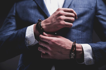 Fashion portrait of young businessman handsome model man dressed in elegant blue suit with accessories on hands posing on gray background in studio. Buttoning his jacket