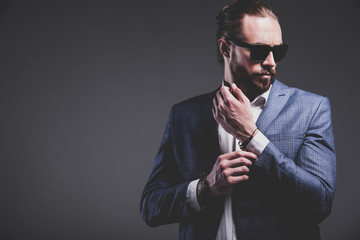 portrait of handsome fashion stylish hipster businessman model dressed in elegant blue suit posing on gray background in studio. Buttoning his jacket