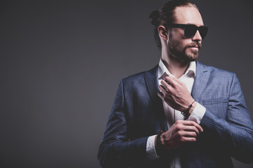 portrait of handsome fashion stylish hipster businessman model dressed in elegant blue suit posing on gray background in studio. Buttoning his jacket