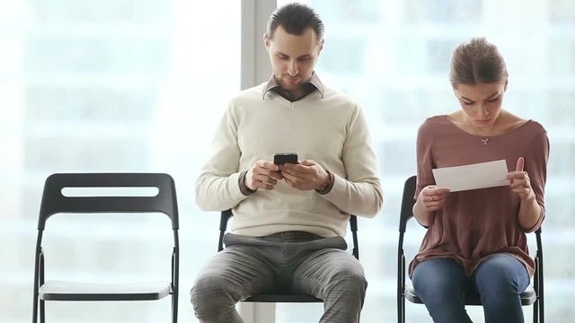 Two young applicants waiting their turn for job interview on chairs in row. Young rejected worried woman running out from office room while confident rivals watching her leave with happy expressions