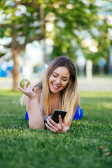 Beautiful attractive young woman eating apple, looking at mobile phone and enjoying outdoors. Healthy lifestyle concept.