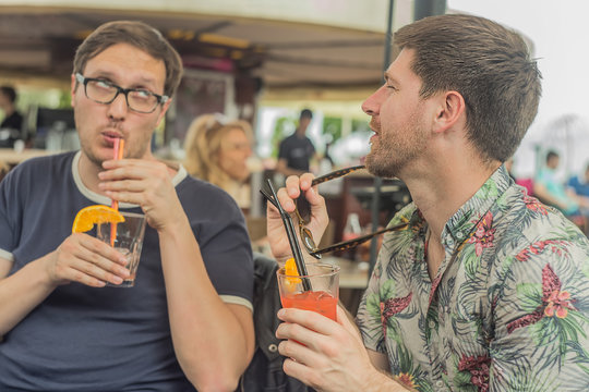 Two young male friends having fun, drinking cocktails and chatting with friends on terrace cafe in town in summer
