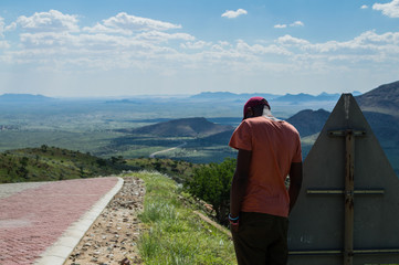 Fototapeta na wymiar Man Standing Next to Sign Post, Lush Mountain Landscape in Namibia