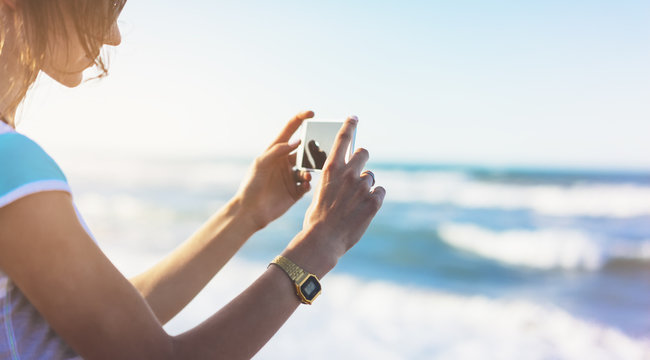 Hipster girl photograph on smart phone gadget in sand coastline, mock up of blank screen. Traveler hold and using in hand mobile on background seascape horizon on blue sun ocean, lifestyle