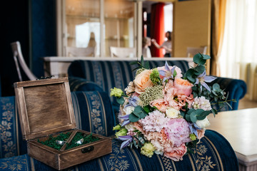 A wedding bouquet of flowers and a wooden box with wedding rings on the couch. The make-up artist paints the bride on the background.  