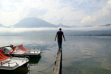 Travel to Sankt-Wolfgang, Austria. The young man are walking on a bridge with the view on the lake near to mountains in the cloudy weather.