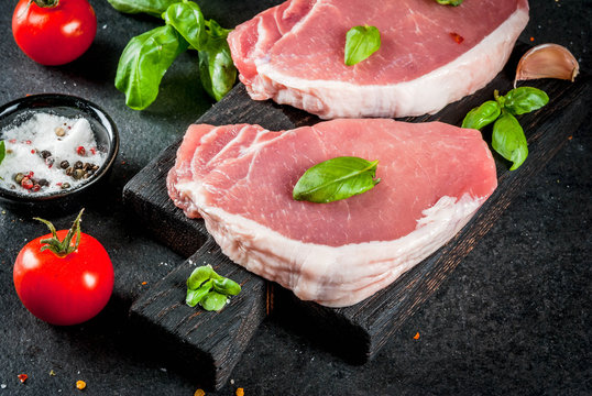 Raw organic meat. Pork steaks, fillets for grilling, baking or frying. On a wooden cutting board, with salt, pepper, basil, tomatoes, garlic. On a gray stone table. Copy space