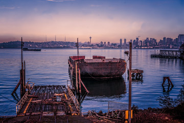 Blue, purple, pink early morning view of shipyard repair area and a rusty barge with Seattle skyline background. 