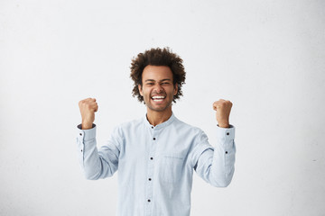 Yes! Waist up studio shot of lucky successful mixed race male student with Afro haircut exclaiming,...