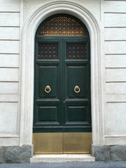 Green double doors with fanlight and brass knockers.  Stone cladding and stained glass windows.  Rome, Italy.