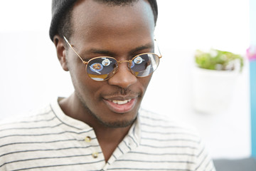 Close up portrait of positive happy young African American man wearing trendy sunglasses and hat smiling in anticipation of tasty breakfast, table with food is reflected on mirror lens of his shades