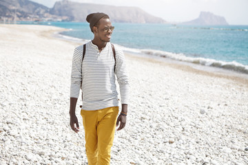 Fototapeta na wymiar Happy young Afro American male tourist dressed stylishly smiling broadly enjoying seascape and sunny summer day, walking on desert pebble beach, exploring coast and admiring beautiful marne views