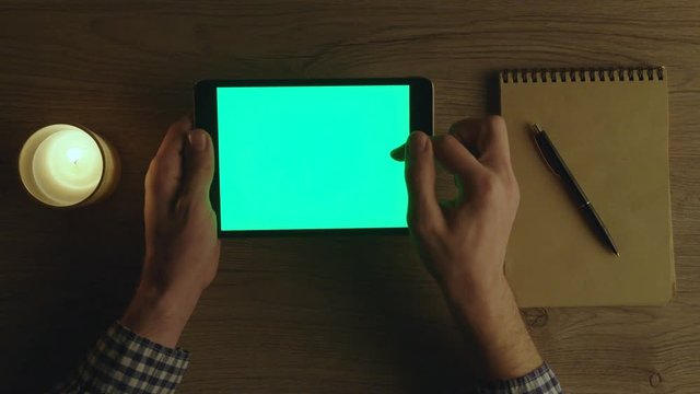 Man using tablet device with green screen at office at night. Chroma key. Table top view. Man scrolling pages, sufring the internet