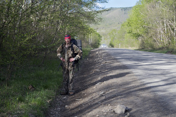 Obraz premium A traveler or a tourist with a large backpack and rug goes along the road near the forest and tries to catch a car (hitch-hiking)