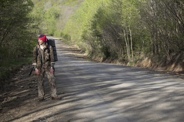 A traveler or a tourist with a large backpack and rug goes along the road near the forest and tries to catch a car (hitch-hiking)