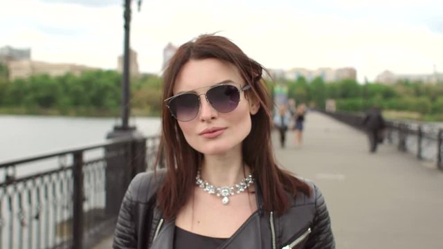 Attractive girl in sunglasses and jewelry walks on a pedestrian bridge. Portrait of a beautiful brunette walking on the bridge.