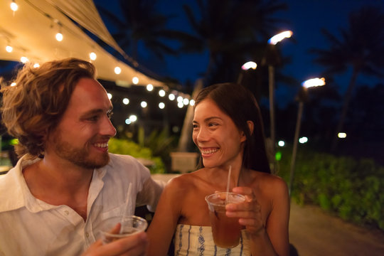 Couple Dating Drinking At Bar On Night Out At Outdoor Restaurant Terrace In Hawaii Vacation Travel. Asian Woman, Man Having Fun Together Toasting Mai Tai Drinks, Hawaiian Cocktail. Luxury Lifestyle.