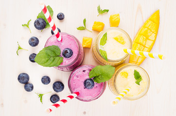 Freshly blended yellow and violet  fruit smoothie in glass jars with straw, mint leaves, mango slices, berry, top view. Soft white wooden board background.