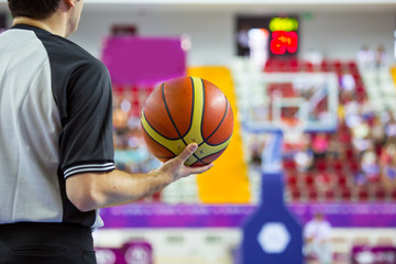 Basketball referee holding a basketball at a game in a crowded sports arena.
