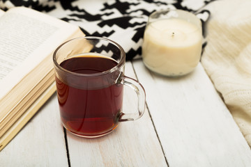 A cup of warm tea, a warm blanket, a candle and a book on a wooden background