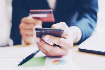 Closeup view of businessman holding credit card in hand and using smartphone,paper documents on the wooden table.Blurred background.Horizontal.