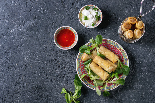Fried spring rolls with red and white sauces, served in traditional china plate and fry basket with fresh green salad over black texture background. Flat lay, space. Asian food