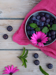 Fresh blueberries in metal bowl,  leaves of mint and pink flowers on white wooden desk.